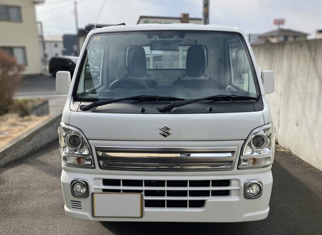 キャリイトラック 買取相場 | 車買取のソコカラ Front view of a white Suzuki van parked on a street, showing the smiling bumper and chrome grille.