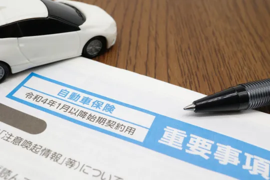 White toy car sits on a wooden desk beside a Japanese auto insurance form with a blue header and a black pen nearby.