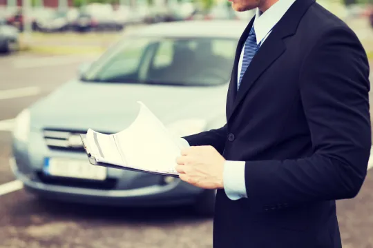 CG車査定 スタッフ お店 | 車買取のソコカラ Man in a dark suit and tie holds scattered documents in front of a parked car outside the frame edge shows the vehicle behind him.
