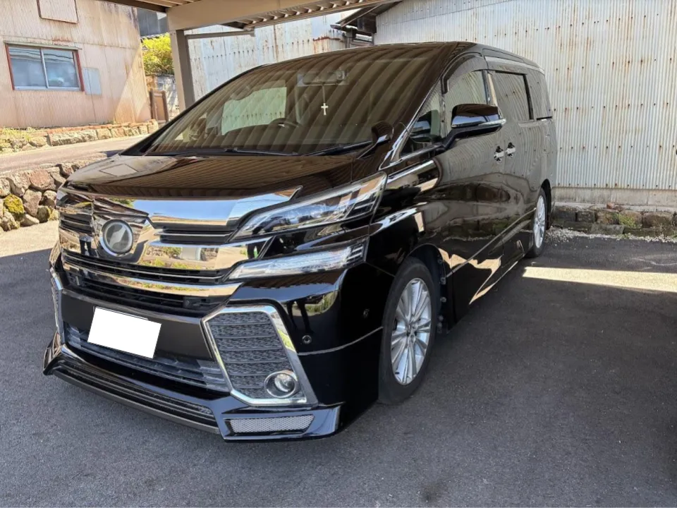 ヴェルファイア買取事例 | 車買取のソコカラ Black minivan with a chrome front grille and fog lights, parked under a carport.