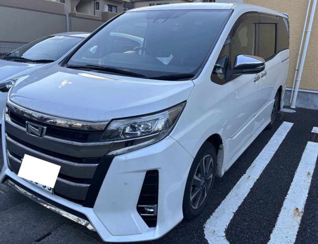 ノア | 車買取のソコカラ White minivan parked in a marked space, viewed from the front-left with a chrome grille and prominent headlights.
