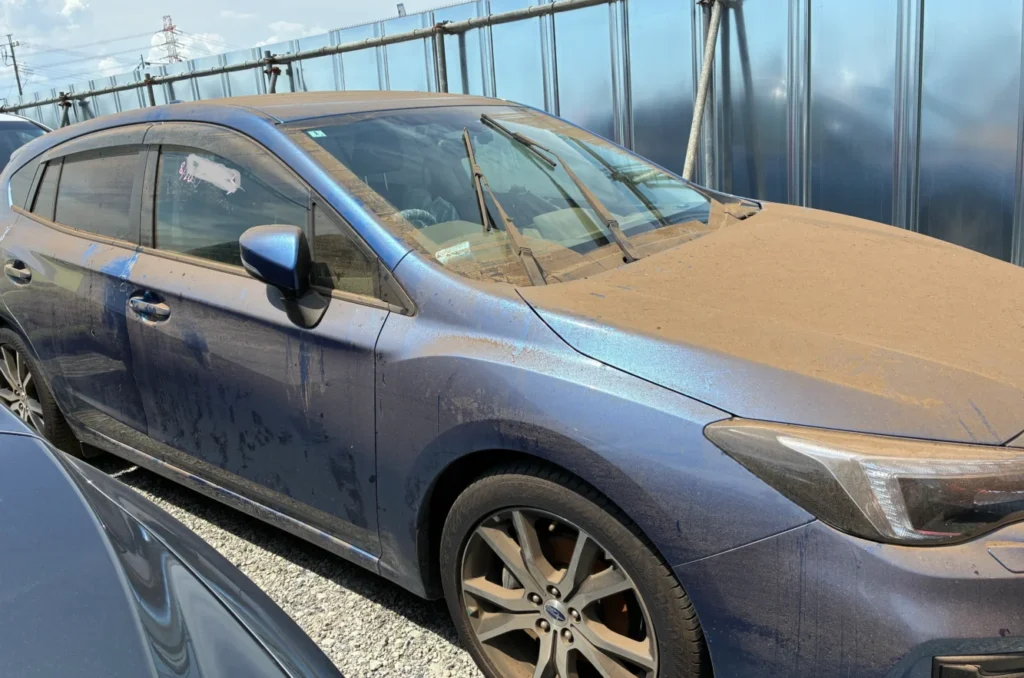 Blue sedan parked in a lot, covered in a thick layer of dust and sand with windshield wipers resting on the windshield.