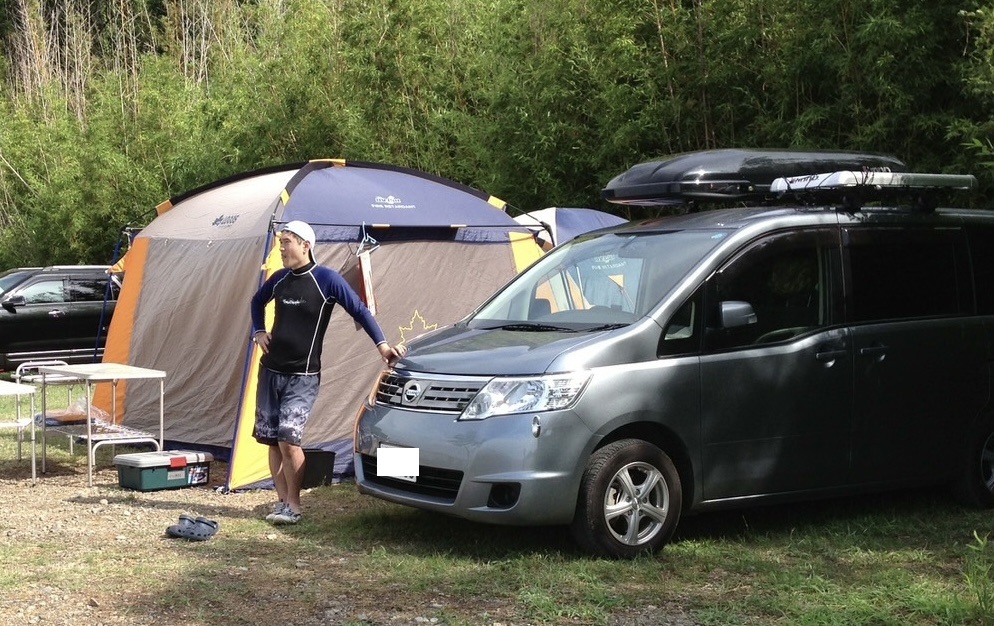 Man stands beside a gray van with a roof rack at a forest campsite, near a large blue-yellow tent.